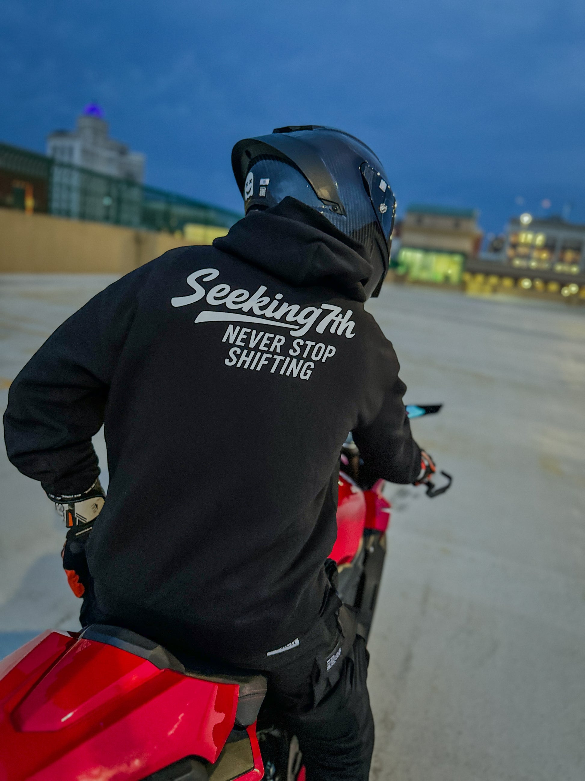 Person wearing a black jacket with 'Seekingth NEVER STOP SHIFTING' text on a red motorcycle at night.