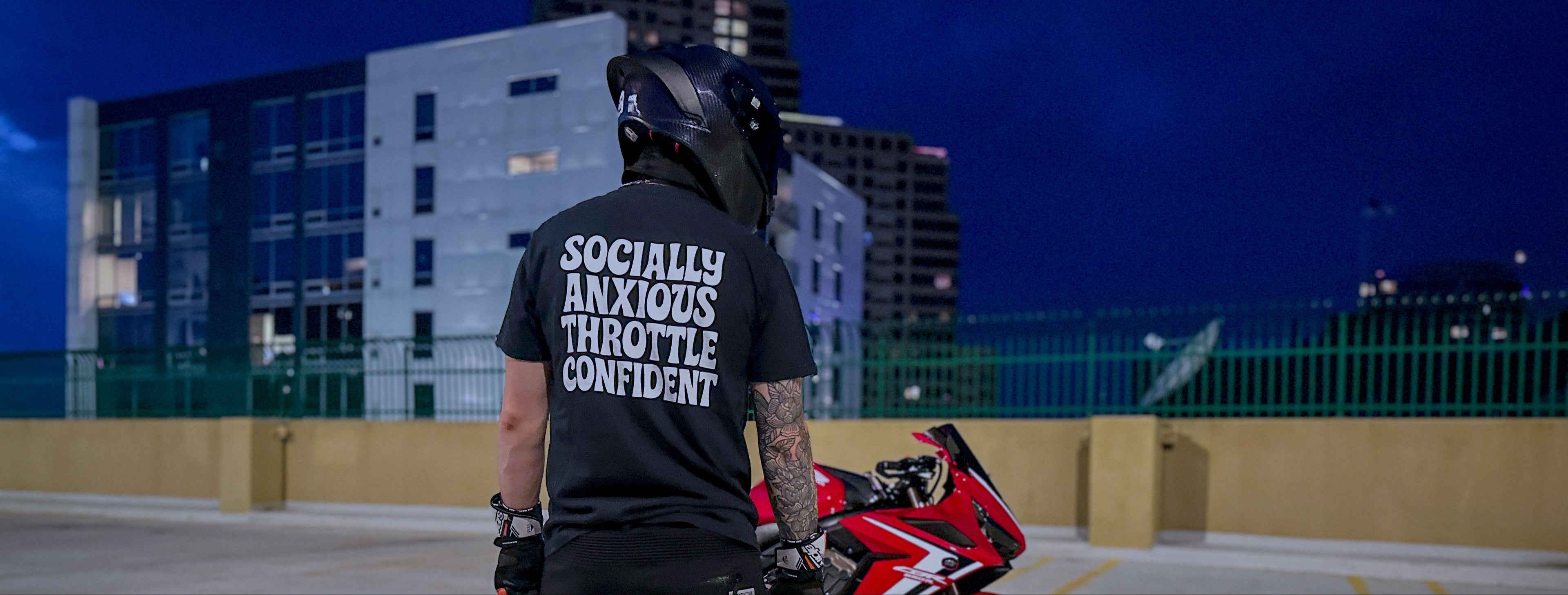 Person wearing a black t-shirt with text standing next to a red motorcycle on a rooftop at night.